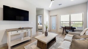 A modern living room with a flat-screen TV, a coffee table, a sofa, and a dining area with a table and chairs. - Landing | Chic 1BD, Game Room, Clubhouse (Mansfield)