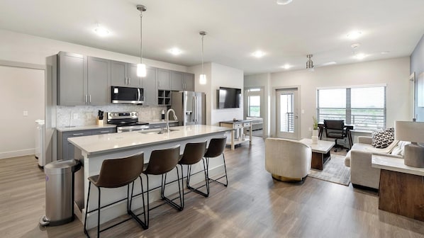 A modern kitchen with a central island, bar stools, and a dining area with a sofa and table. - Landing | Modern 2BD, Clubhouse, Game Room (Mansfield)