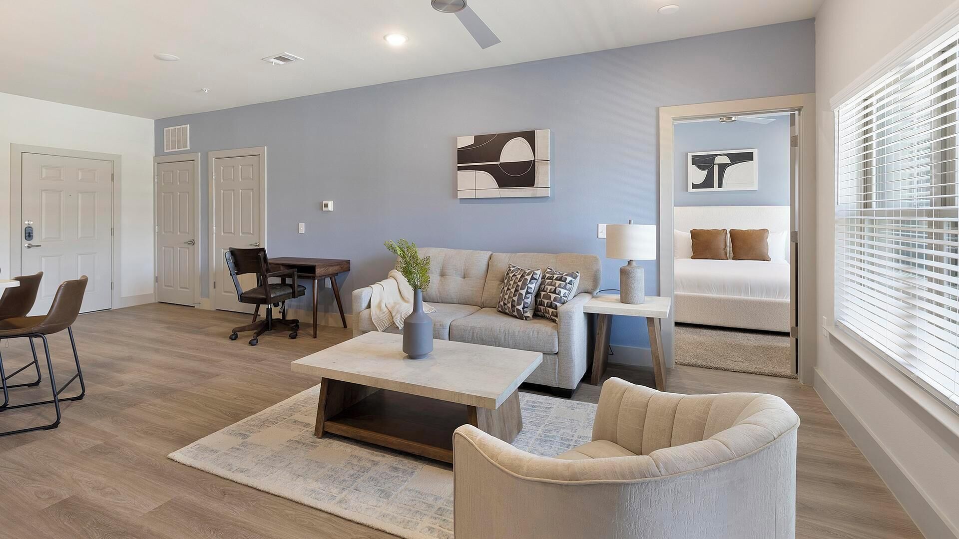 Un salon moderne avec un canapé, une table basse et des fauteuils, un coin repas avec une table et des chaises, et une chambre à coucher avec un lit et des stores aux fenêtres.