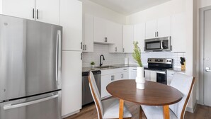 A modern kitchen with stainless steel appliances, white cabinetry, a round wooden dining table, and a potted plant. - Landing | Spectacular 1BD, Gym (Houston)