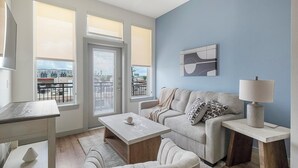 A modern living room with a sofa, coffee table, and lamp. There are large windows and a view of buildings outside. - Landing | Spectacular 1BD, Gym (Houston)