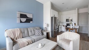 A modern living room with a sofa, a coffee table, and a chair. There is a kitchen area with cabinets, a refrigerator, and a microwave. - Landing | Chic 1BD, Gym (Houston)