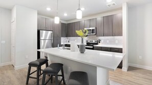 A modern kitchen with dark wood cabinets, stainless steel appliances, a white island with bar stools, and pendant lighting. - Landing | Modern 1BD, Gym, Game Room (Fort Worth)