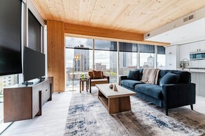 A modern living room with a large TV, wooden ceiling, and a city view. - Landing | Beautiful 3BD, Pool, Gym (Milwaukee)