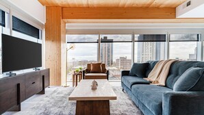 A modern living room with a large TV, a sofa, a coffee table, and a view of the city through large windows. - Landing | Amazing 3BD, Pool, Game Room (Milwaukee)
