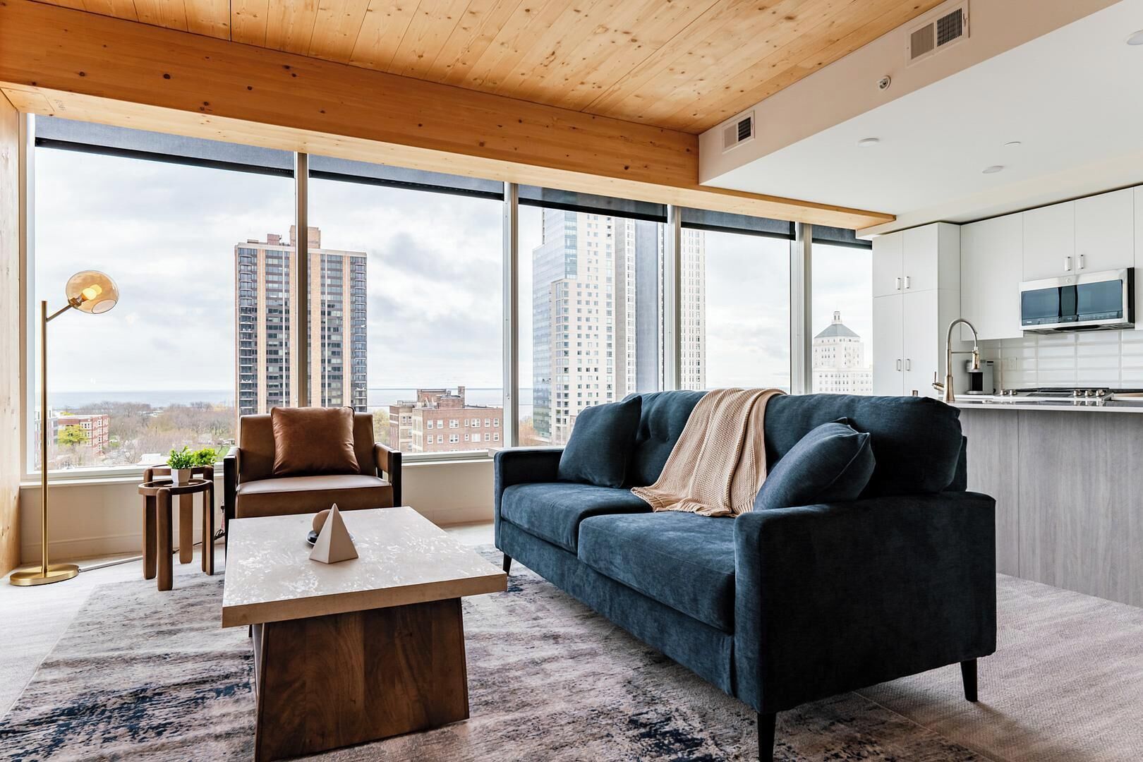 A modern living room with a sofa, coffee table, and a view of tall buildings through large windows.