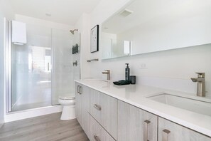 A modern bathroom with a glass shower enclosure, a white toilet, a large mirror, and a wooden vanity. - Landing | Chic 2BD, Gym, Game Room (Milwaukee)
