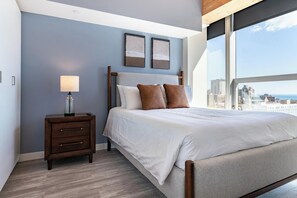 A bedroom with a bed, a nightstand, a lamp, and a city view through large windows. - Landing | Chic 2BD, Gym, Game Room (Milwaukee)