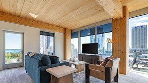 Un salon moderne avec un canapé, une table basse et un téléviseur. De grandes fenêtres offrent une vue sur la ville.