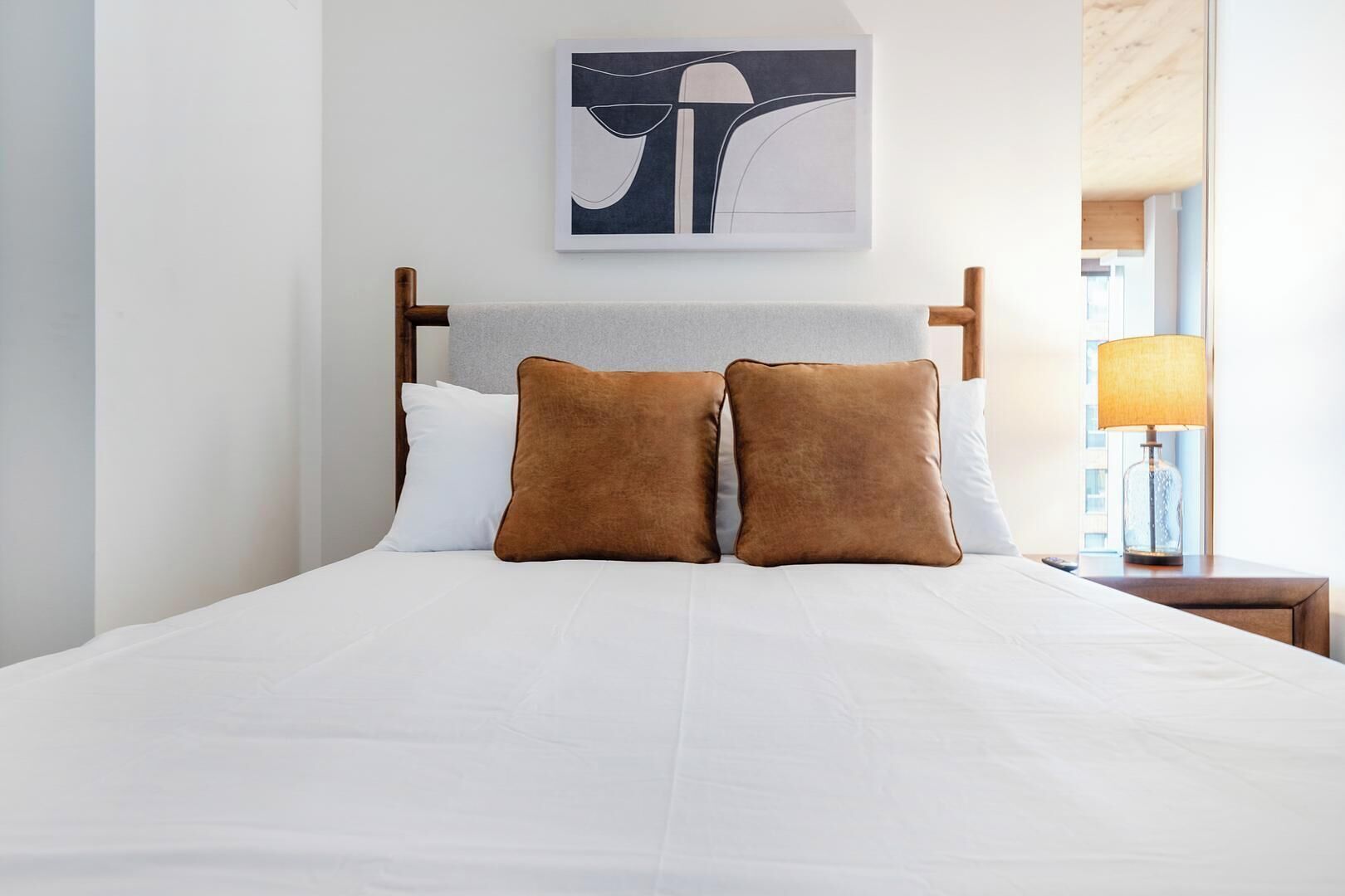 A neatly made bed with white bedding and two brown pillows, a wooden headboard, and a framed abstract artwork above.