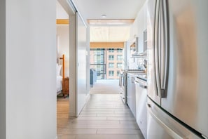 A modern kitchen with stainless steel appliances, a wooden floor, and a view of a building through large windows. - Landing | Stylish 1BD, Game Room, Gym (Milwaukee)