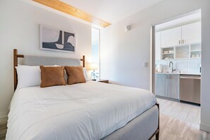 Een slaapkamer met een bed, nachtkastje, lamp, schilderij en uitzicht op een keukenhoek.