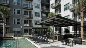 Ein Poolbereich mit Liegestühlen, ein überdachter Grillplatz und ein Gebäude mit mehreren Balkonen.