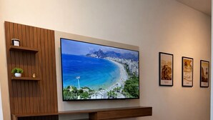 A bedroom with a bed, a mounted TV, a wooden shelf with a plant, and framed pictures on the wall. - New Studio • Sea view • 2 minutes with your feet in the sand (Rio de Janeiro)