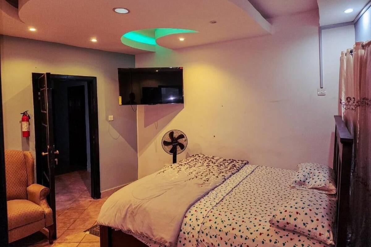 Un dormitorio con cama, una silla, televisor montado en la pared, ventilador de techo y una puerta que da a otra habitación.