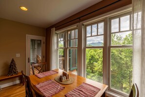 A dining area with a wooden table, chairs, and a view of a forest through large windows. - Gateway to Grandfather | Community Pools, Mountain View, Fitness Rooms, and More (Boone)