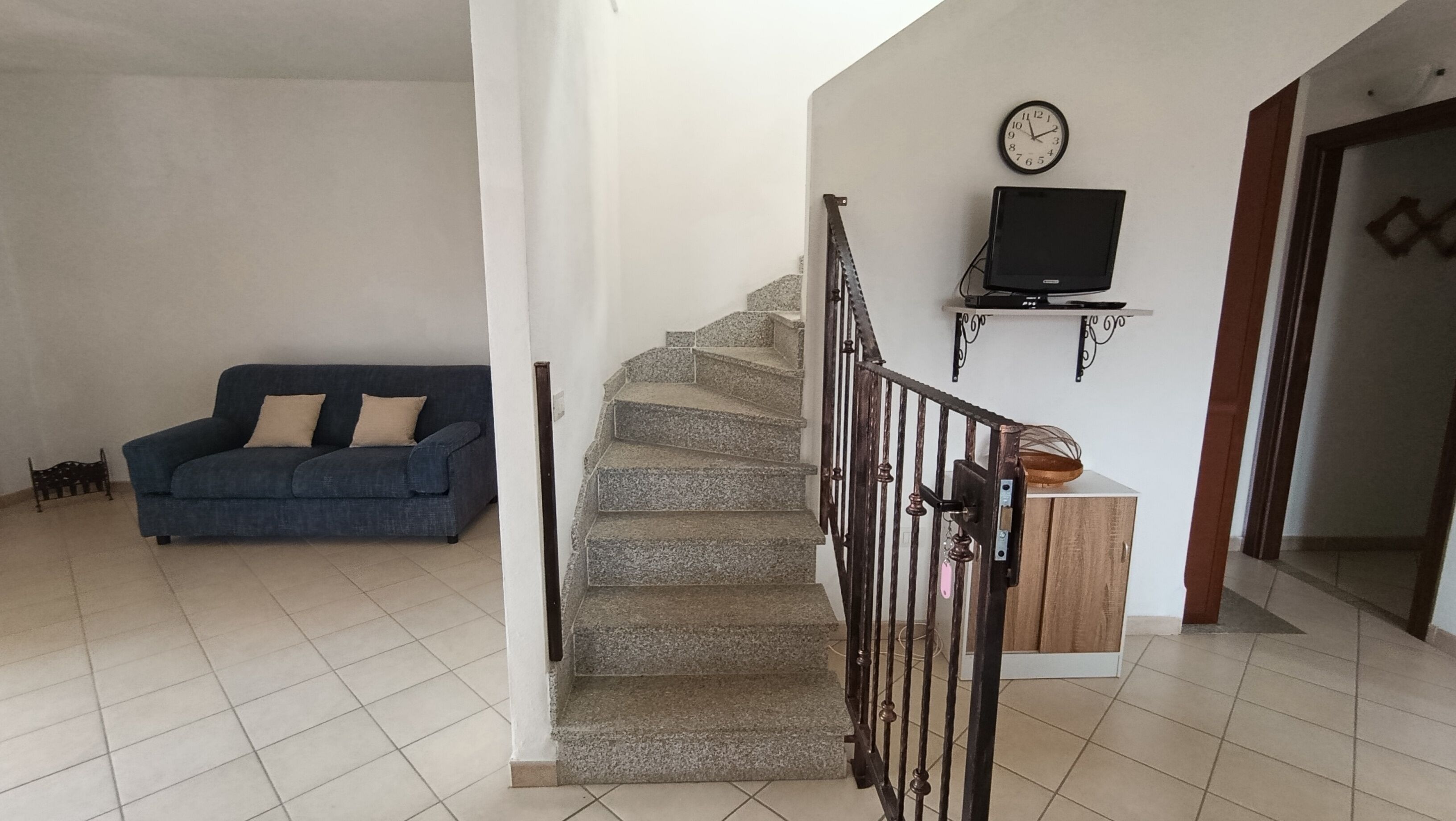 Una escalera con barandilla de madera, un sofá azul con cojines beige y un televisor fijado a la pared.