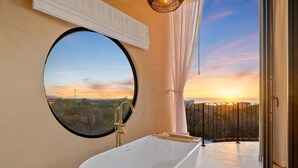 Un baño moderno con una bañera independiente, un ventanal redondo y un balcón con vistas al atardecer.