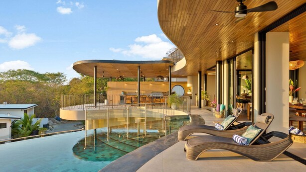 Un área de piscina moderna al aire libre con sillones de descanso, techo de madera y vistas del paisaje circundante.