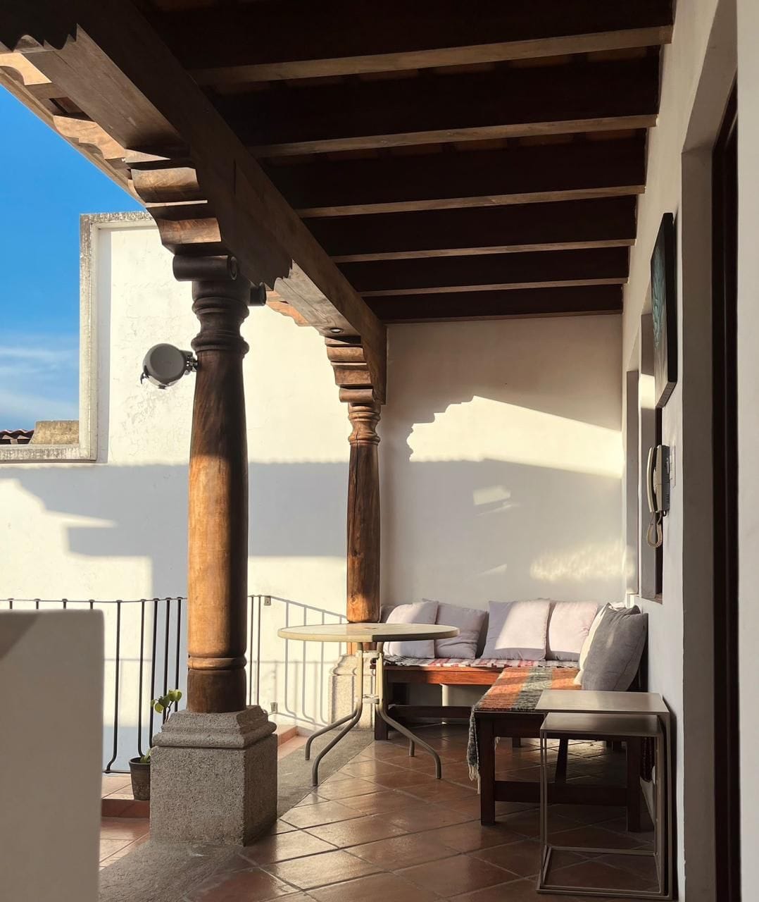 Un balcon agrémenté d’une pergola en bois, un coin salon avec des coussins, une petite table et une plante en pot.