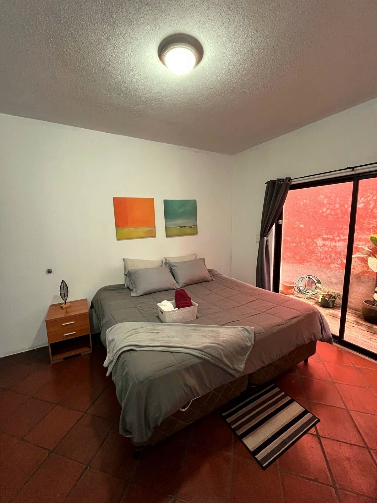 Ein Schlafzimmer mit einem Bett, einem Nachttisch, einem Fenster mit Vorhängen und zwei an der Wand hängenden Bildern.