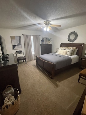 A bedroom with a bed, a ceiling fan, a dresser, a chair, and a desk.
