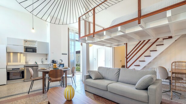 A modern kitchen with built-in appliances, a dining area with a wooden table and chairs, a grey sofa, and a staircase leading to an upper level. - The Happy House - Welkeys (Saint-Georges-d'Oléron)