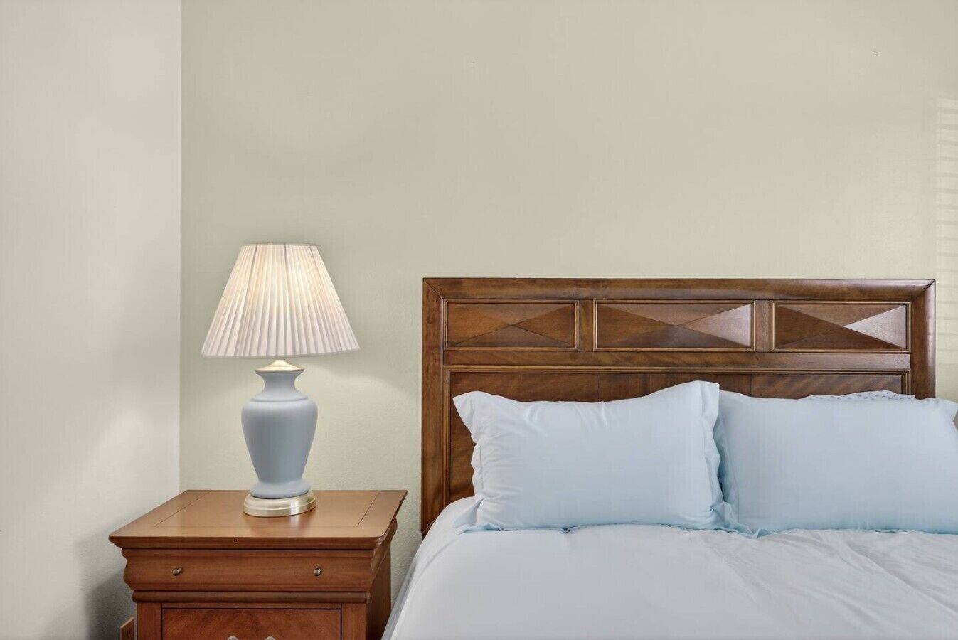 A bedroom with a wooden headboard, a bedside table with a lamp, and two pillows on the bed.
