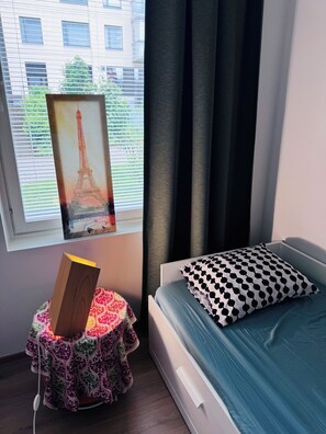 Ein Schlafzimmer mit einem Bett, einem Nachttisch mit einem Buch, einer gemusterten Tischdecke, einem Fenster mit Blick auf Gebäude und Vorhängen.
