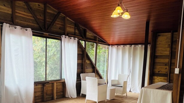 Ein Zimmer einer Holzhütte mit einem Bett, zwei Stühlen, einem kleinen Tisch und großen Fenstern mit Vorhängen.