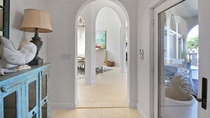 A hallway with a white door, a blue cabinet with a lamp, and an archway leading to a room with a painting and a plant. - Waterfront Neo Mediterranean Home On Casey Key - Tidewater Manor (Nokomis)