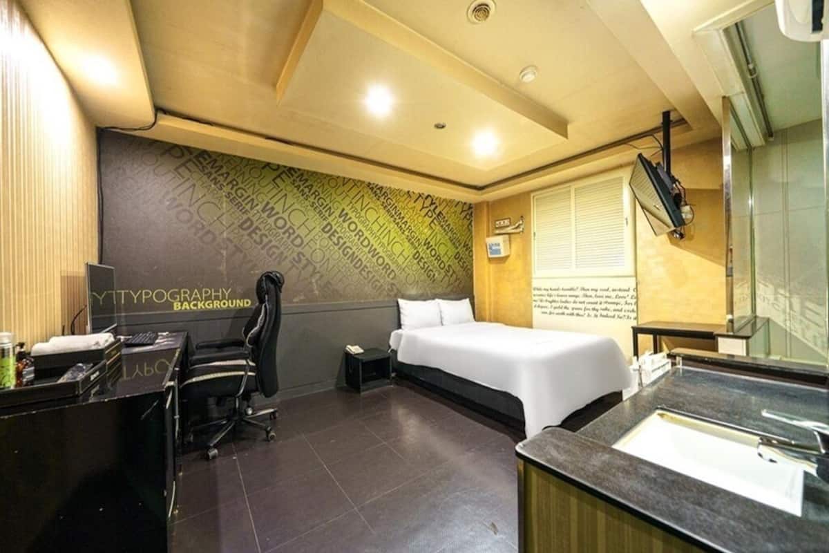 A modern hotel room with a bed, a desk, a chair, a bathroom with a sink, and a wall with typography.