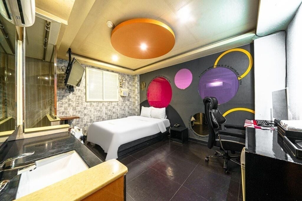 A modern hotel room with a large bed, a desk, a chair, a bathroom with a sink, and a shower.