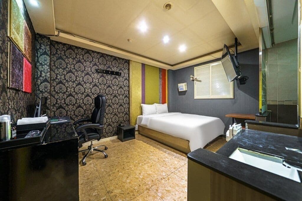 A hotel room with a bed, desk, chair, and a bathroom with a sink and mirror.