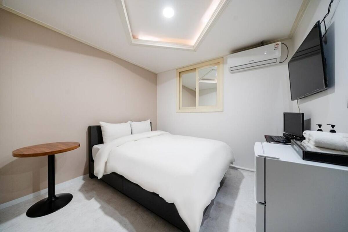 A modern hotel room with a bed, a small round table, a desk with a computer, a TV, and an air conditioning unit.