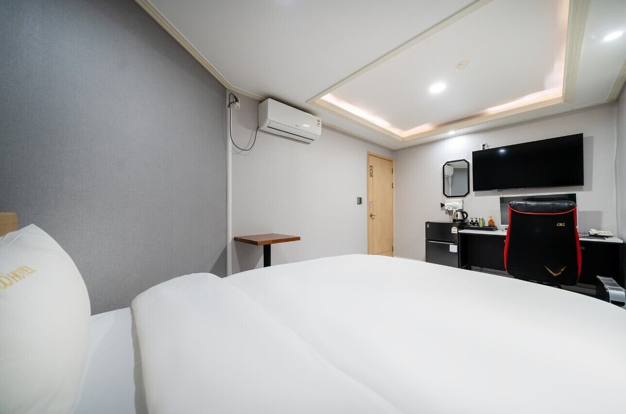 A hotel room with a bed, a desk, a chair, a TV, and an air conditioning unit.