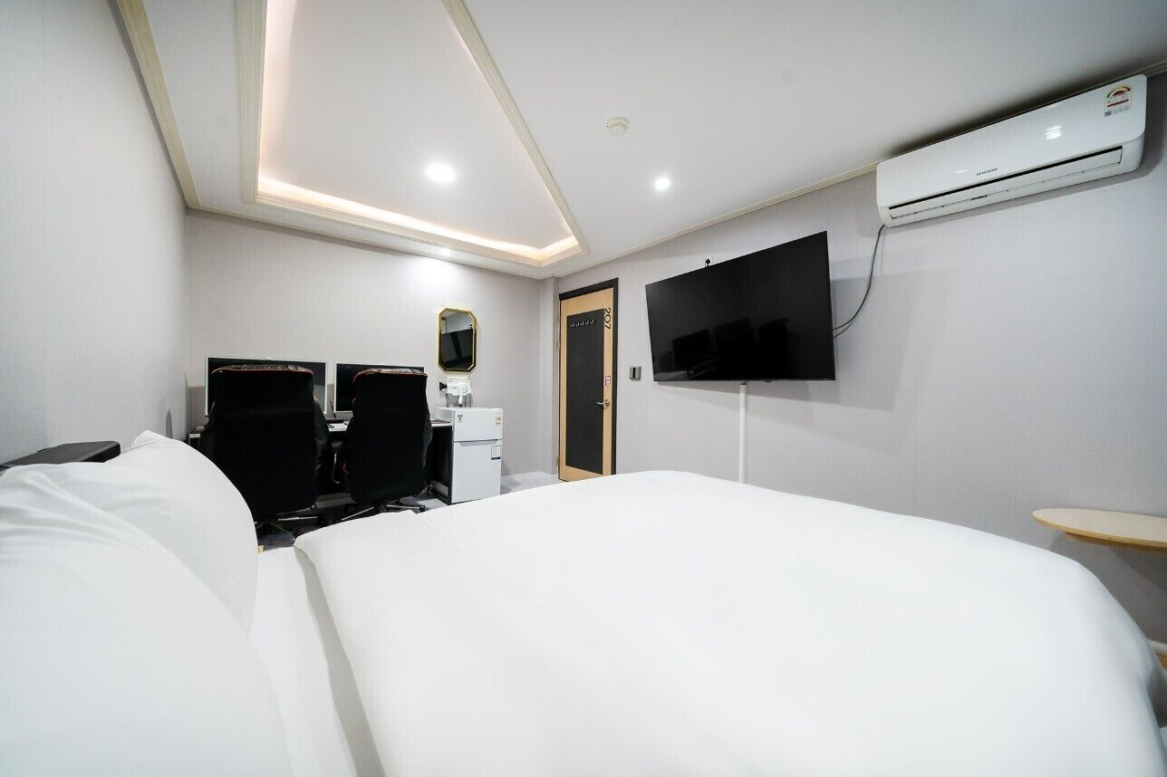 A hotel room with a bed, a desk with a chair, a flat-screen TV, an air conditioning unit, and a door.