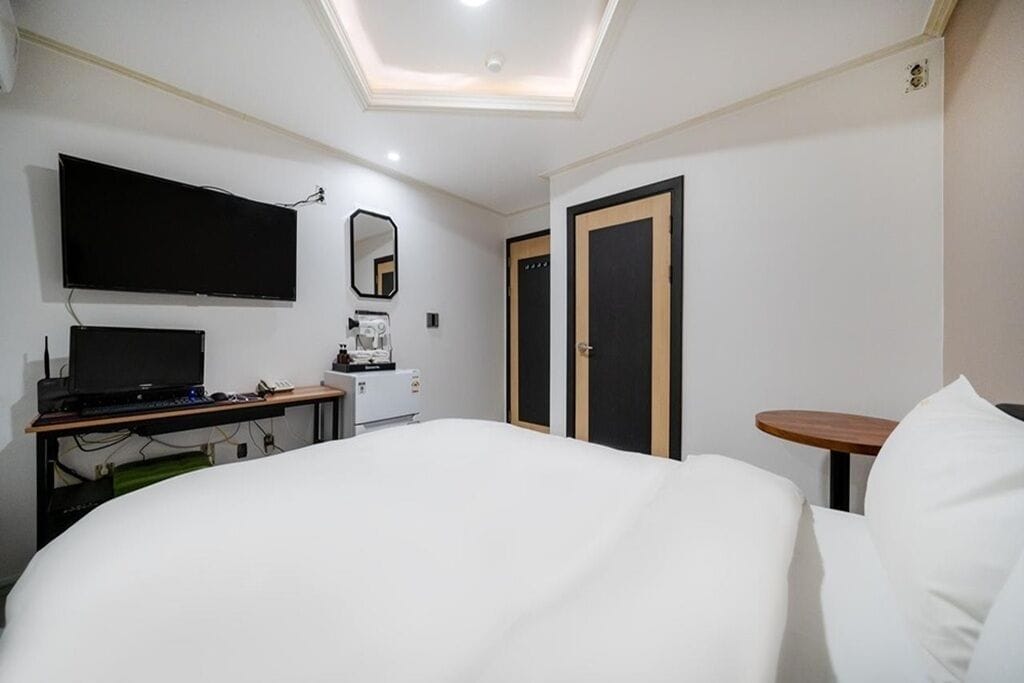 A hotel room with a bed, a desk with a computer, a TV, a mirror, and two doors.