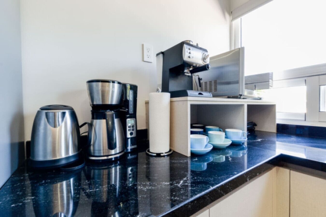La cuisine est équipée d’une machine à café, d’une bouilloire et d’un four à micro-ondes. On peut y voir des tasses et des soucoupes bleues posées sur le comptoir.