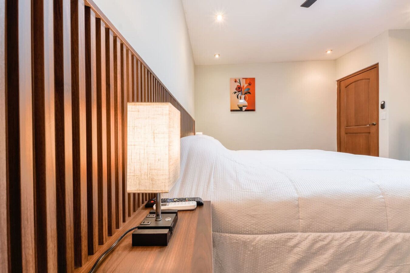 A hotel room with a bed, a bedside table with a lamp, a remote control, and a painting on the wall.