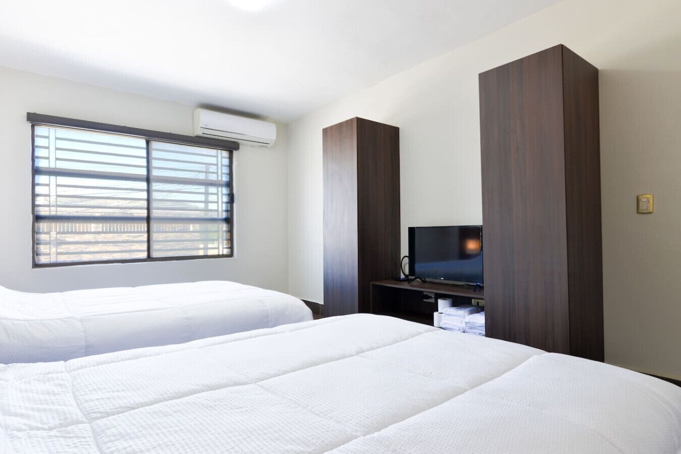 A hotel room with two beds, a TV on a wooden stand, and an air conditioning unit on the wall.