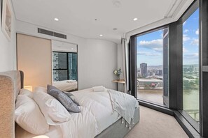 Ein modernes Hotelzimmer mit einem großen Bett, einem Spiegel, einem kleinen Tisch mit einer Vase und Blick auf die Stadt durch große Fenster.