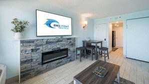 A living room with a stone fireplace, a flat-screen TV, a dining table with chairs, and a potted plant. - Decorated Oceanfront  Condo with Amazing Heated Pool (Myrtle Beach)