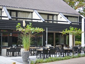 Ein Außenbereich mit Tischen und Stühlen, ein großer Pflanzkübel mit Pflanzen und ein Gebäude mit einem Ziegeldach und Fenstern.