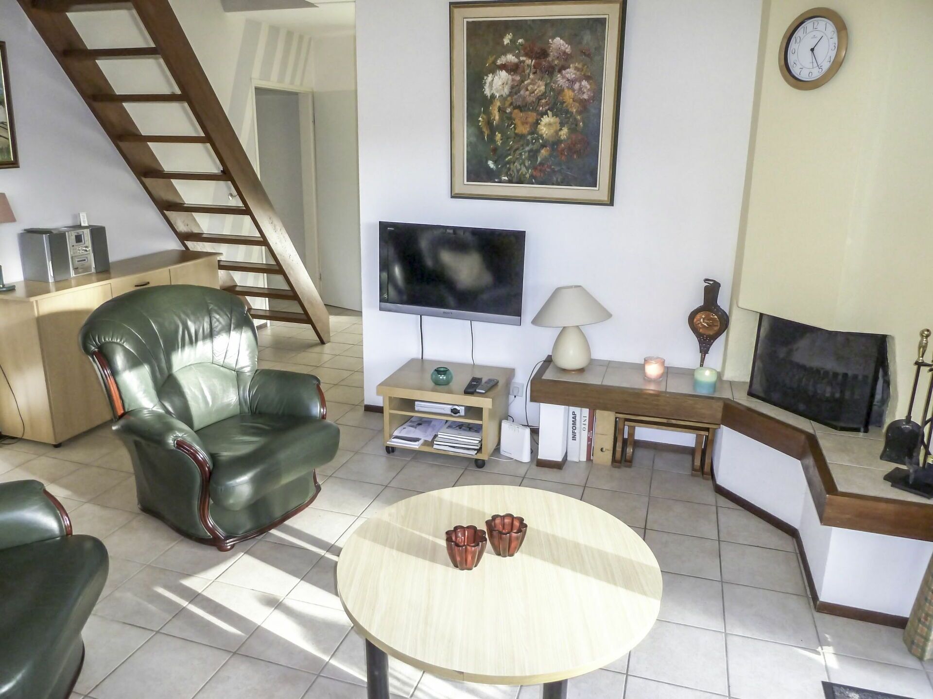 Uma sala de estar com sofá de couro verde, mesa de centro redonda, televisão, quadro, relógio e lareira.