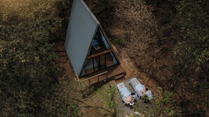 Uma cabana com telhado triangular rodeada por uma floresta densa, com um deck de madeira e duas pessoas deitadas no chão.