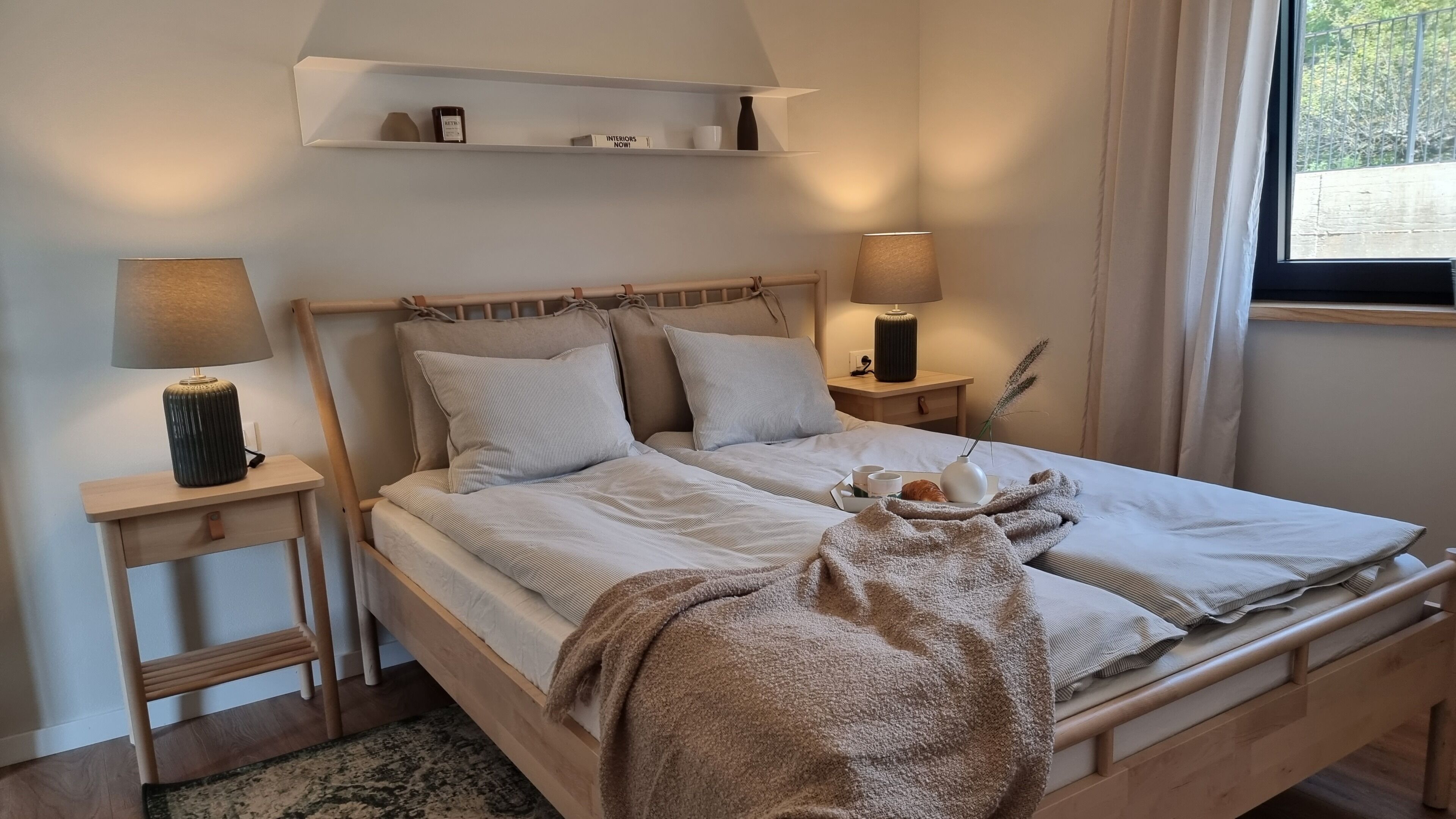 A neatly made bed with pillows, a bedside table with a lamp, a shelf above the bed, and a window with curtains.