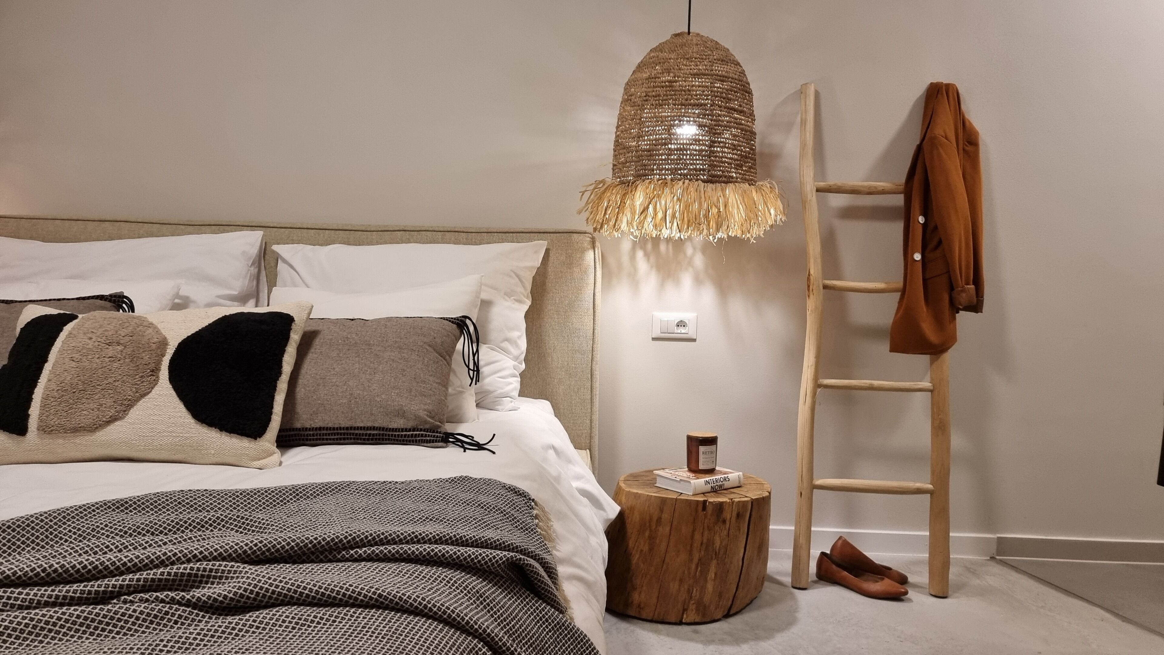 Un dormitorio con cama, almohadas, una lámpara colgante, una escalera de madera y una pequeña mesa redonda con un libro.