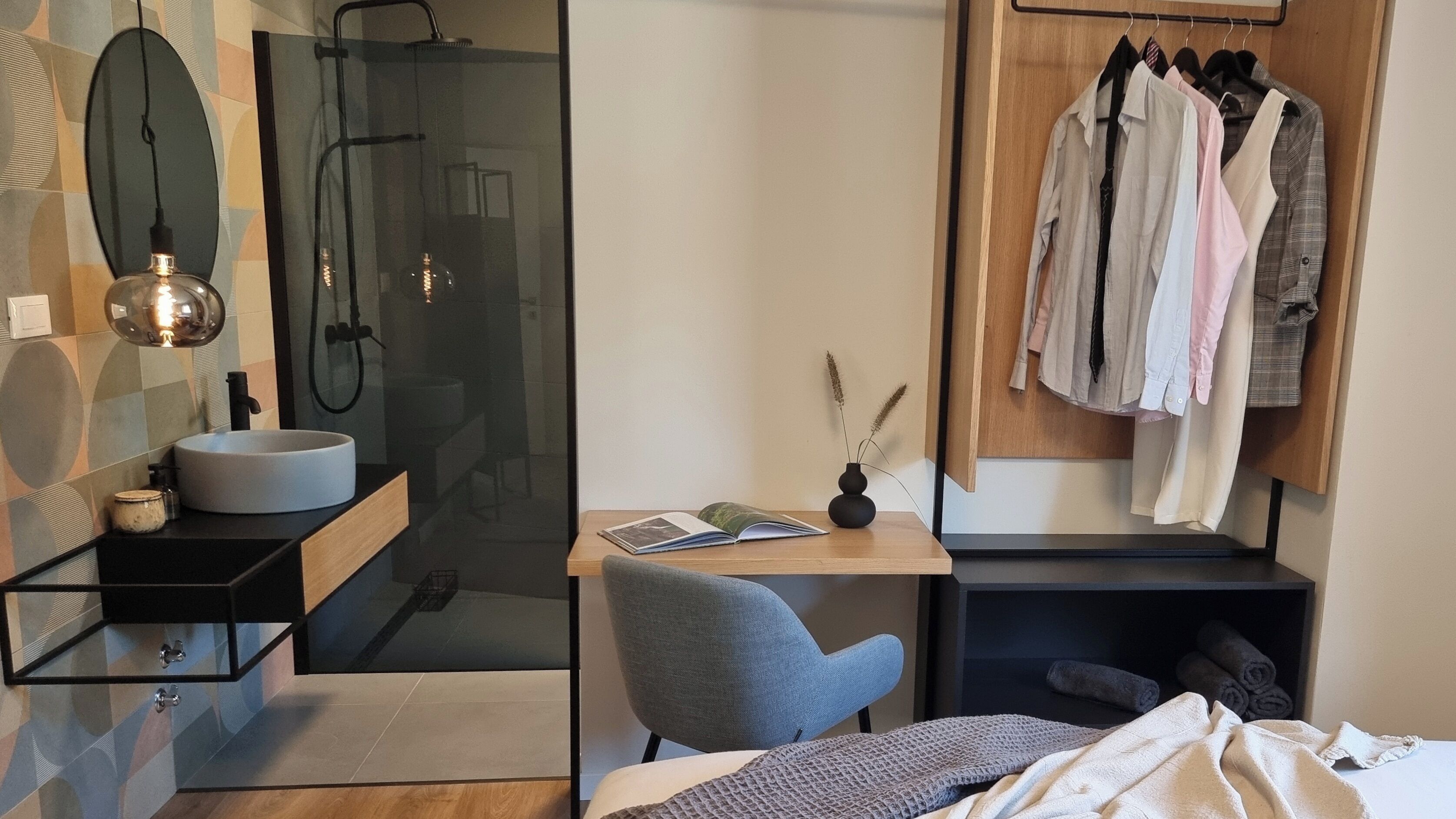 A modern bedroom with a bed, a bedside table with a book, a chair, a desk with a laptop, a mirror, and a wardrobe.
