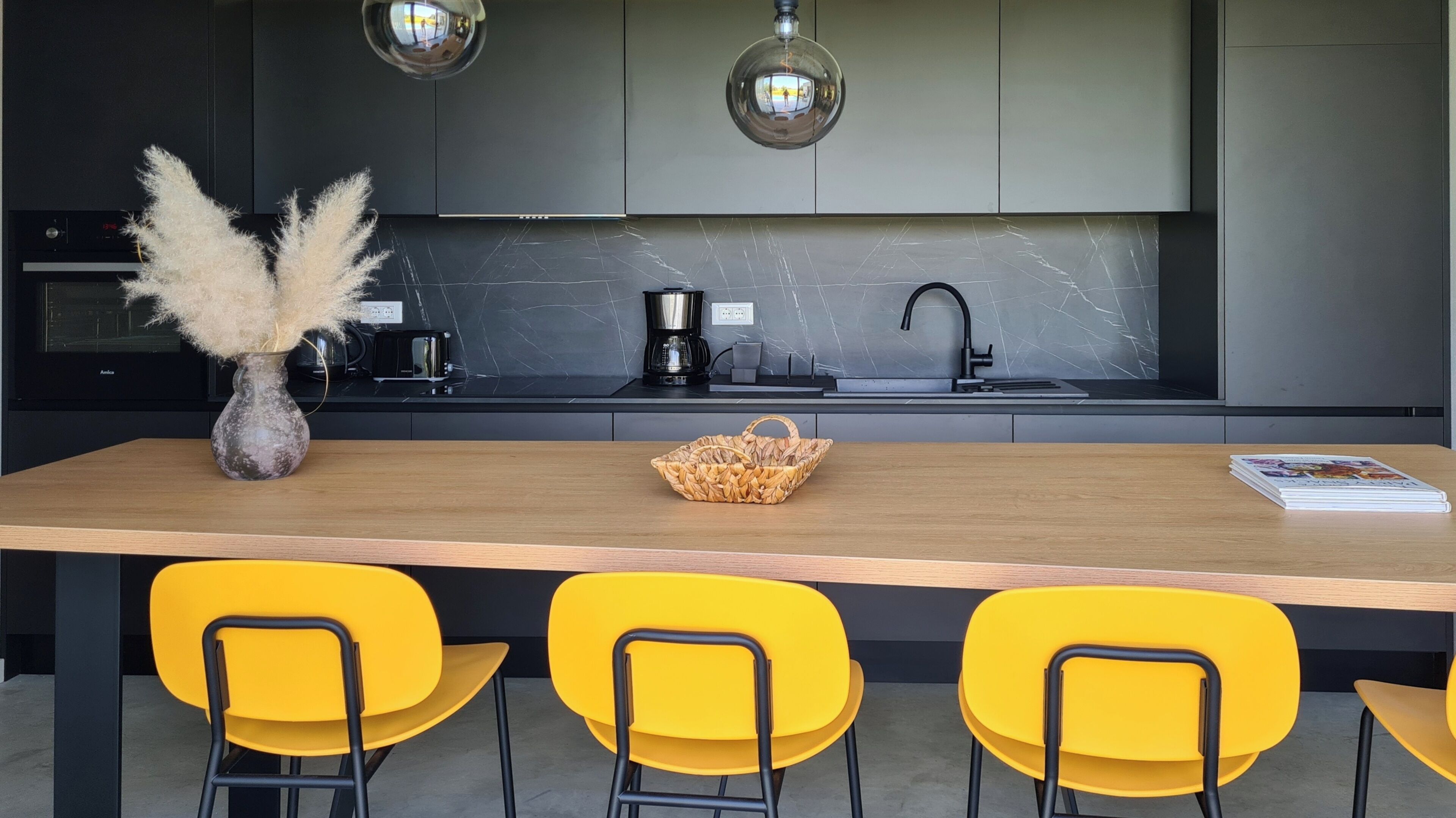 A modern kitchen with a marble countertop, a coffee maker, and a sink. There are three yellow chairs and a woven basket on the table.